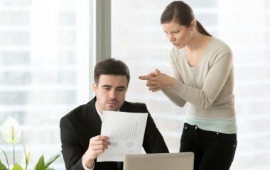 Angry woman aiming with finer gun at head of her hateful boss who attentively reading financial report at desk. Intrigues of office colleagues, bad coworkers relations, hidden negative among employees